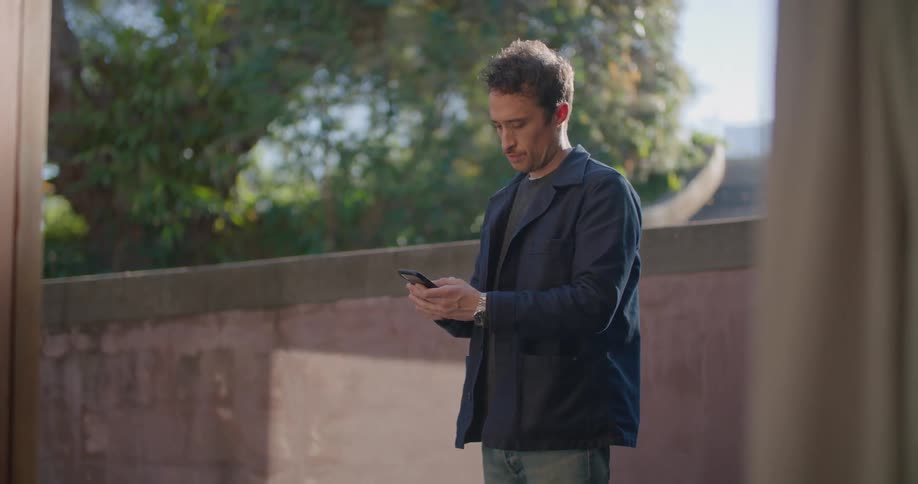 A man standing using a smartphone with a wall a driveway trees the sky and clouds in the background