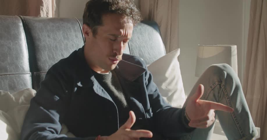 A man sitting on a bed looking at a laptop with a wall and a lamp in the background