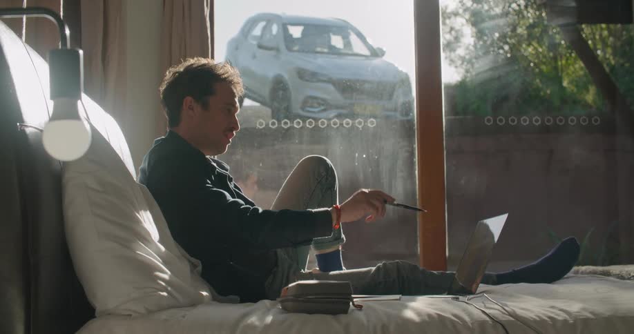 A man reclines on his bed with his laptop nearby as he has a conversation