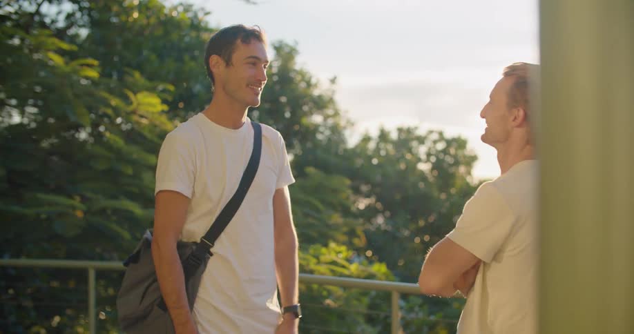 A pair of men greet one another with a hug outdoors before one man leads the other out of frame