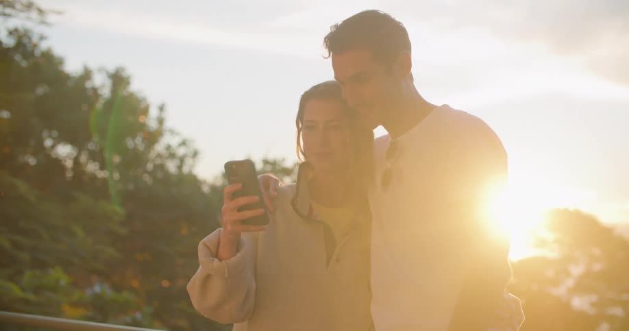 Man with white t shirt and blonde woman in oversized sweatshirt hold each other and uses smartphone at sunset