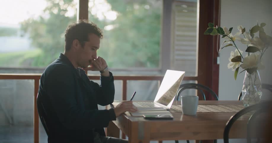 Man sits at table contemplating laptop near window showing traffic