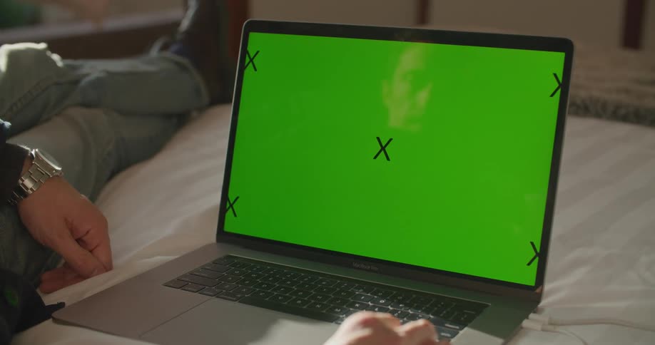 A man looks at the screen of a laptop while resting on a bed