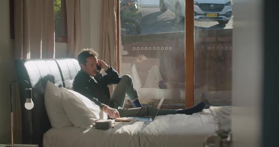 A man in a room with a smart phone sitting on a bed next to a laptop