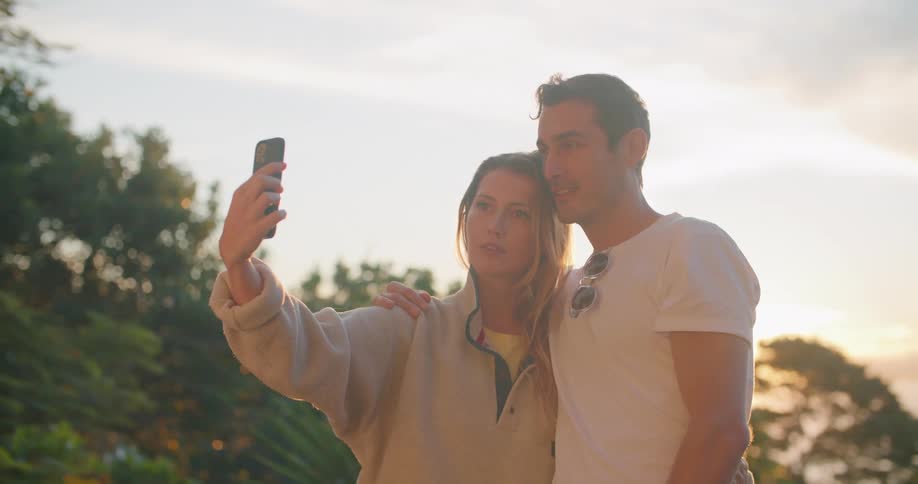A woman smiles and the man embracing her makes funny faces as the woman takes a selfie with her smartphone