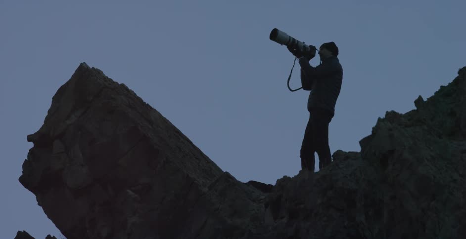 A man on the edge of a cliff takes a photograph at sunset