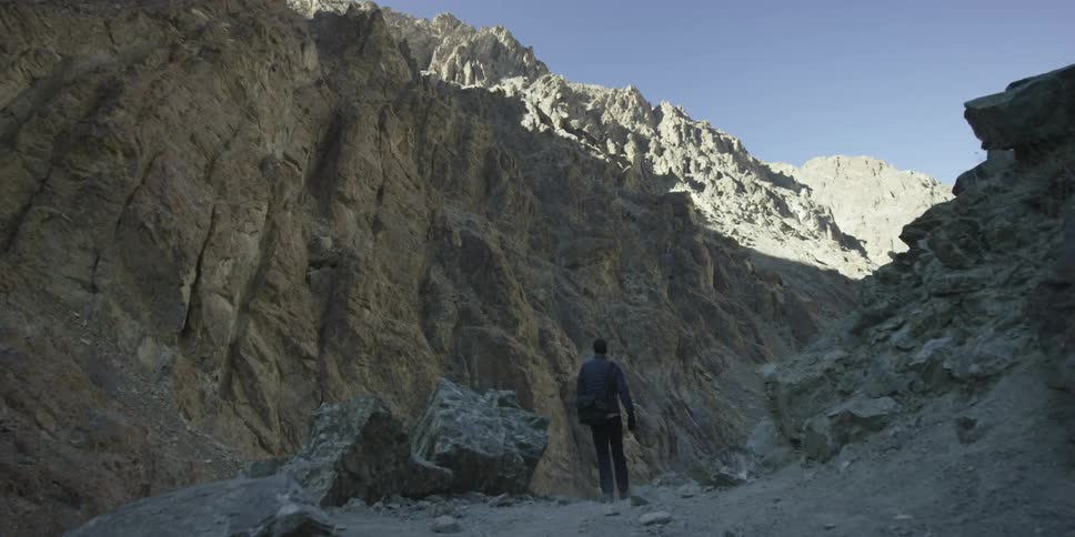 A man walks on a dirt path beneath a tall mountain cliff during the day