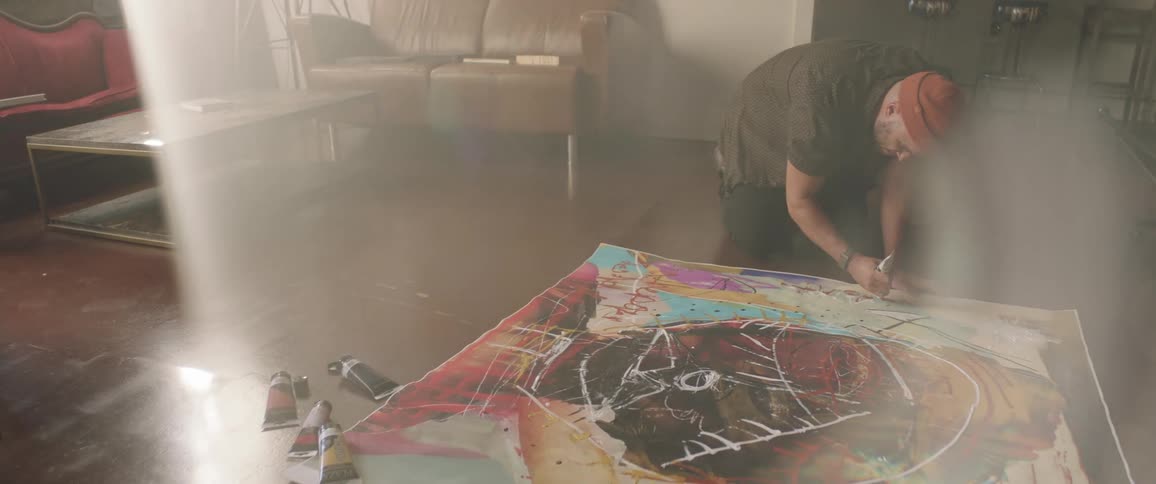 Man kneels on the floor and applies paint from a tube onto an art canvas