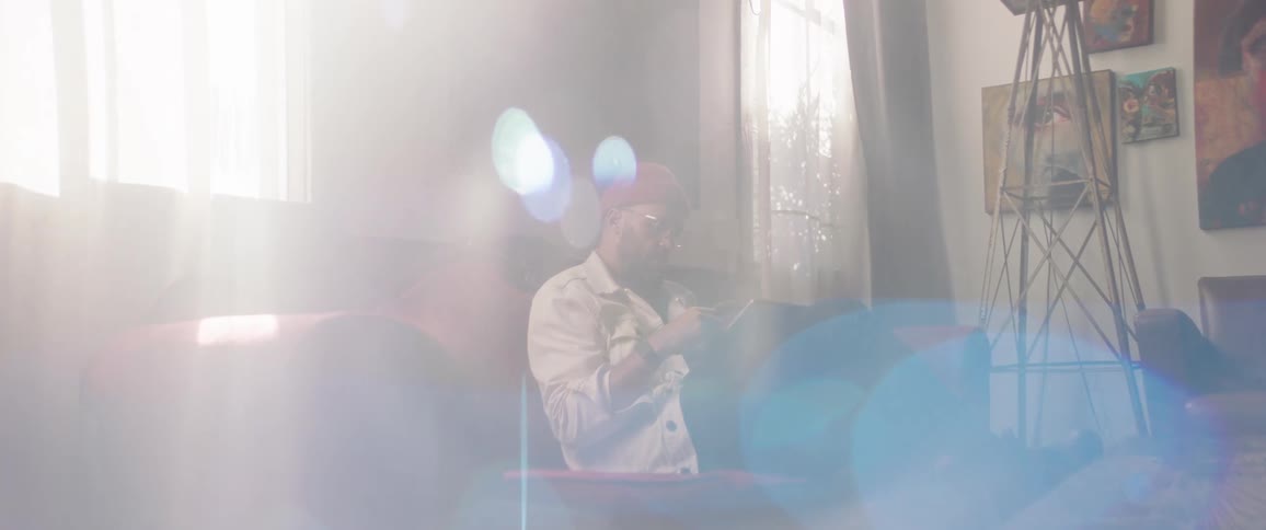 Man with a beard a red woolly hat a white jacket sits on a red couch and uses a tablet in the art studio