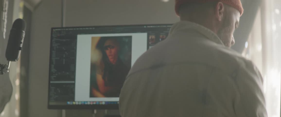 A man standing looking at a screen indoors next to a computer screen with editing software and a picture of a woman