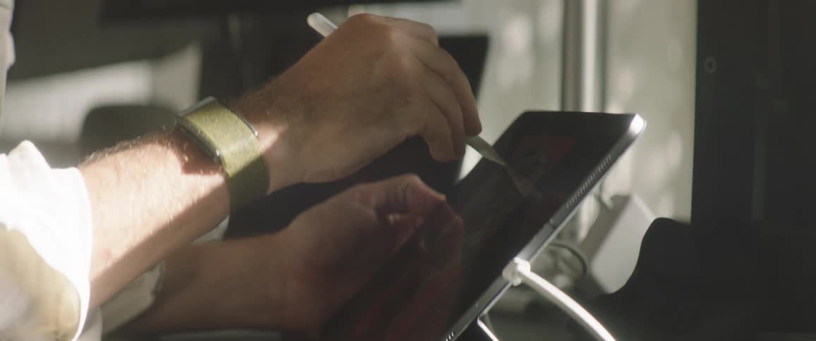 A man sketches on a tablet on top of a table next to a computer screen during the day