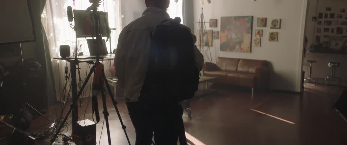Man carrying backpack walks down hallway into photo studio stopping at table set with various computers and screens