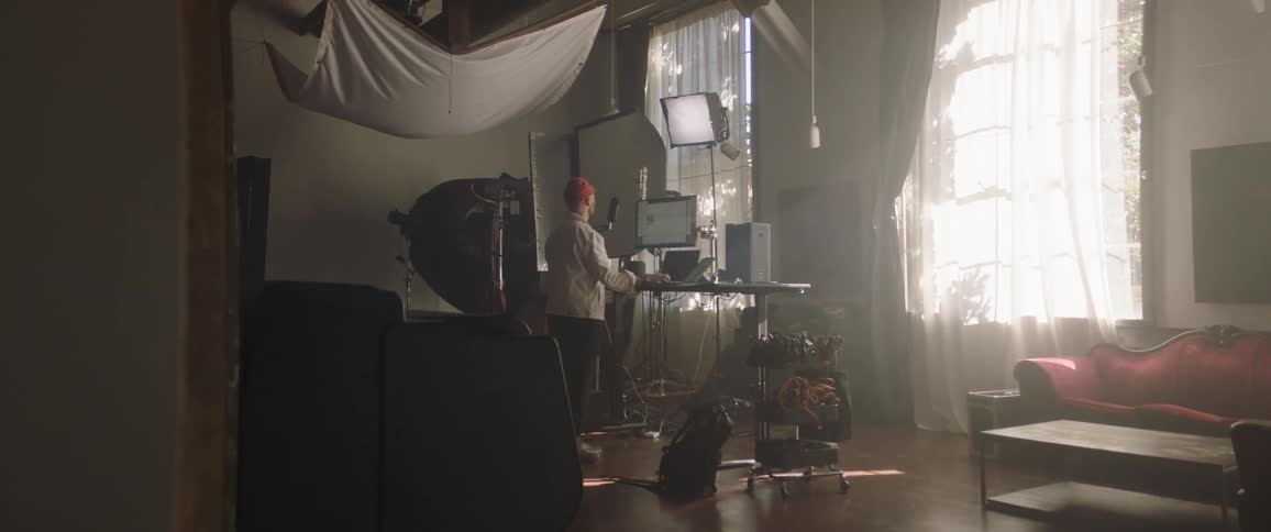 A man with a hat in a room standing at a desk with a computer screen and a microphone