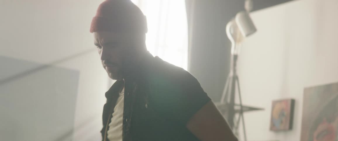 A man in a red beanie walks around a room with a number of paintings on the walls