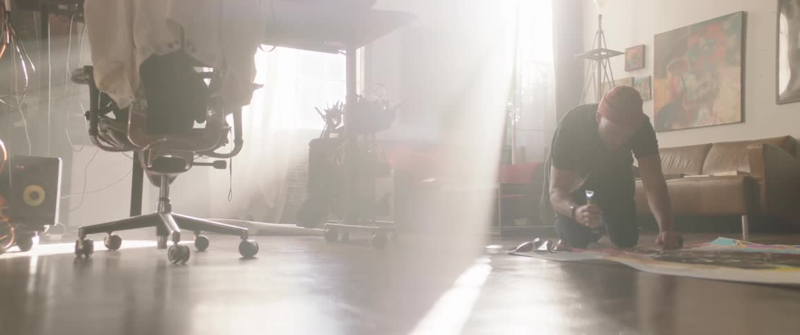 A man uses a tube of paint on a canvas while kneeling on the floor