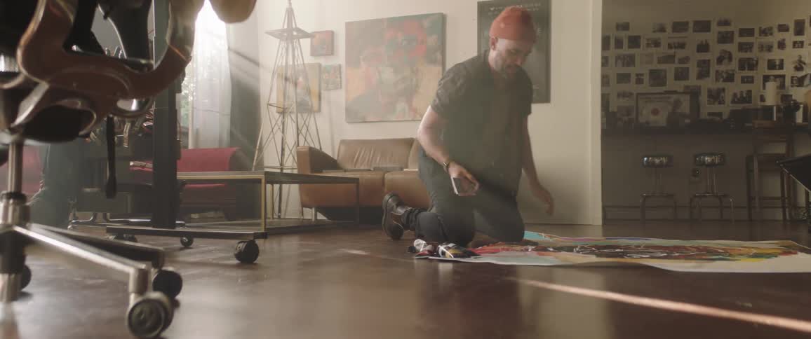 Artist kneels on floor applying paint to canvas in art studio near large printer