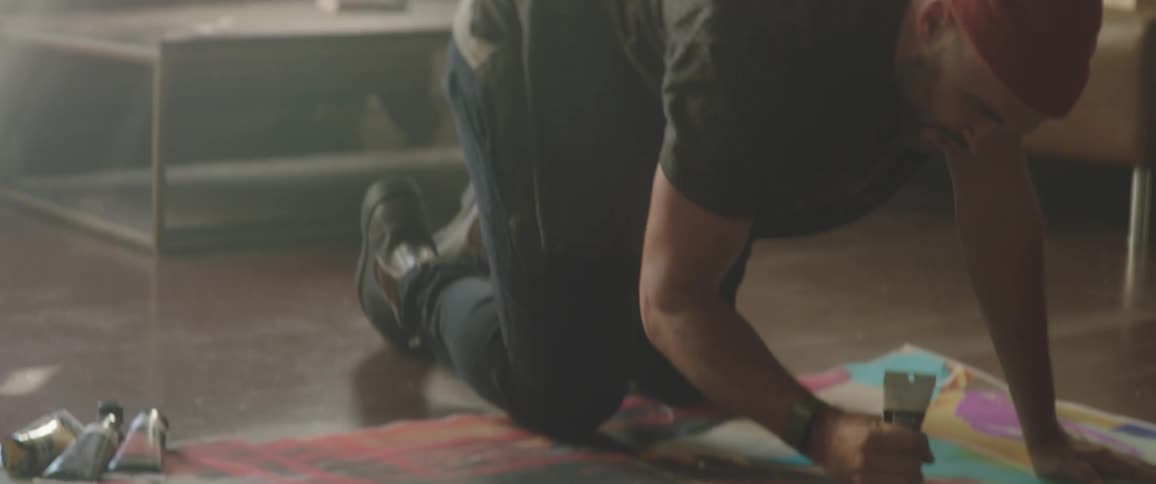 A male painter kneels over a piece of canvas fabric on the floor and applies paint to it