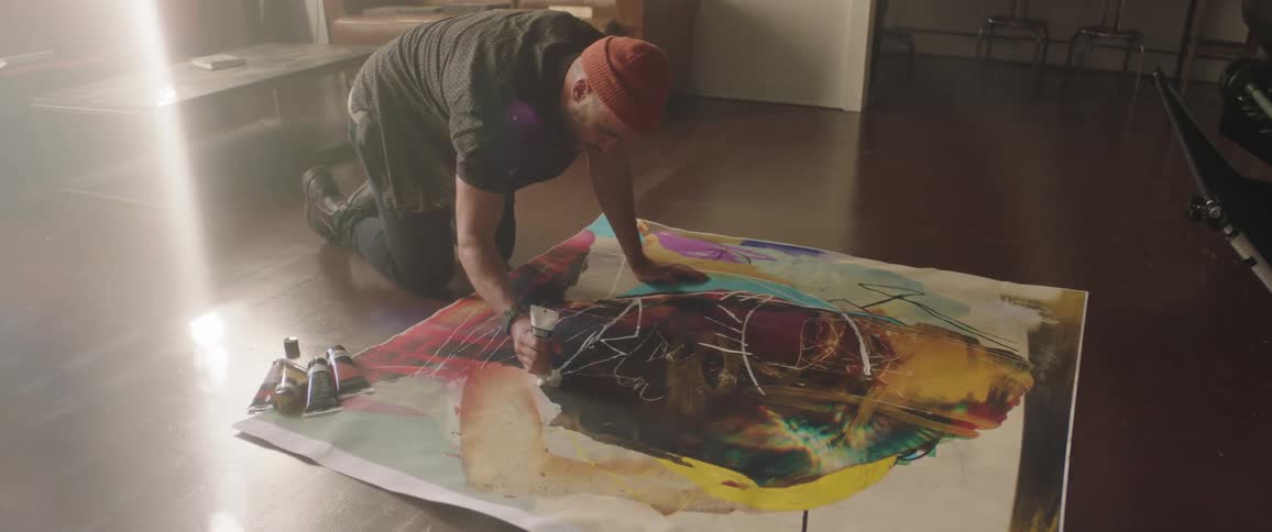 Artists squeezes paint onto painting in progress while kneeling on floor