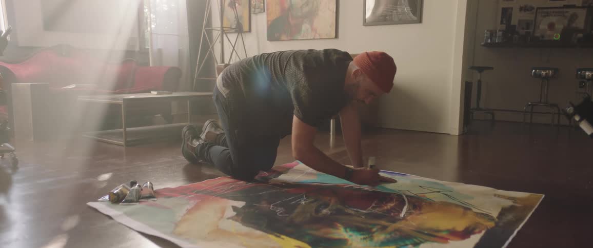 An artist paints on a large canvas lying on the floor with a tube of paint
