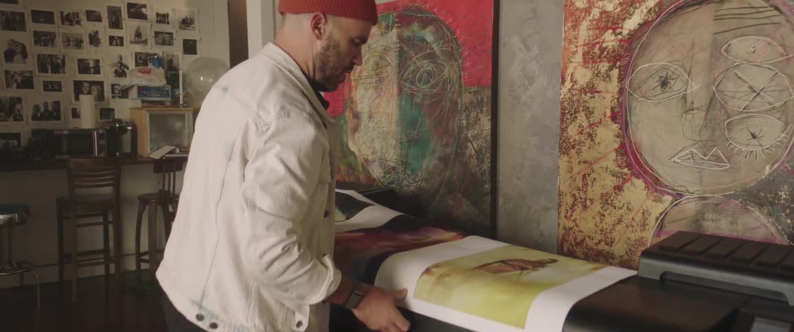 Man with a beard a red woolly hat a white jacket looks at large printed pictures in the art studio