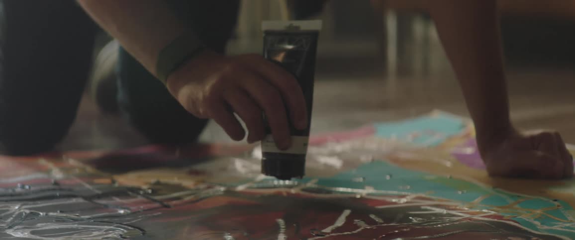 A man with a hat in a room applying paint to a canvas on the floor