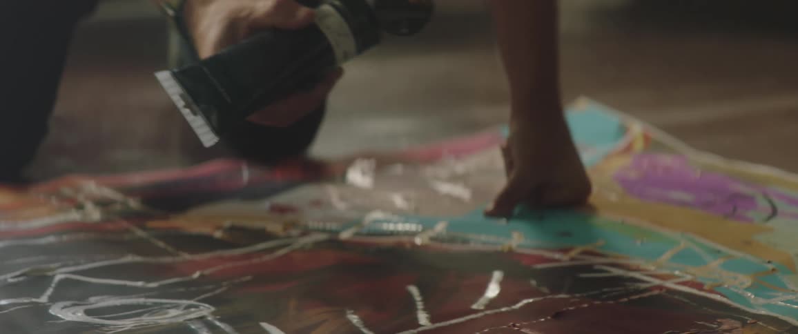 A man uses a tube of paint to paint on a canvas lying on the floor