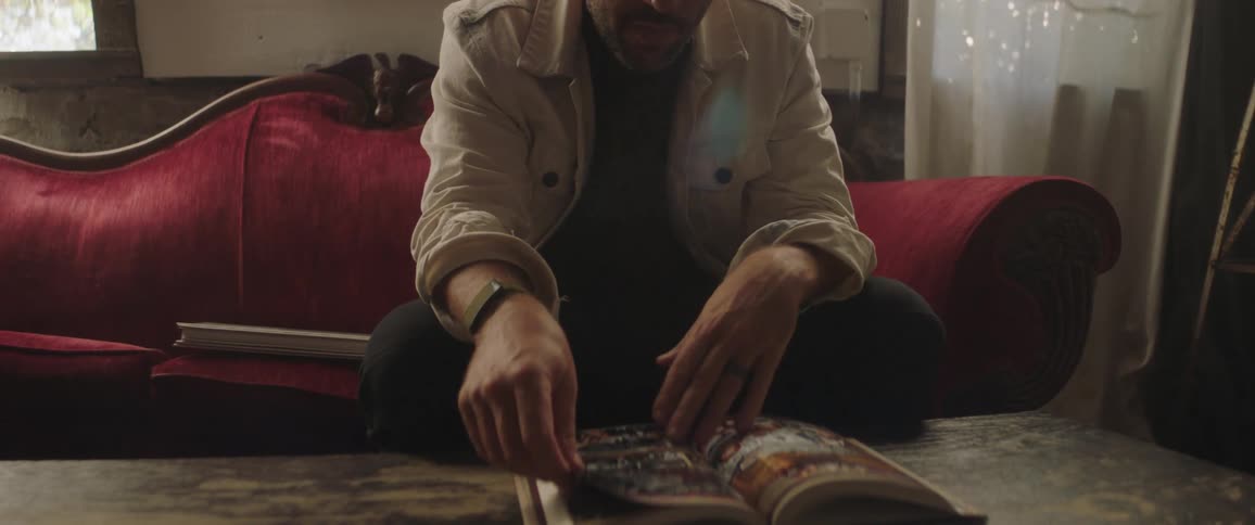 A man sits on a vintage red couch and flips through a thick magazine