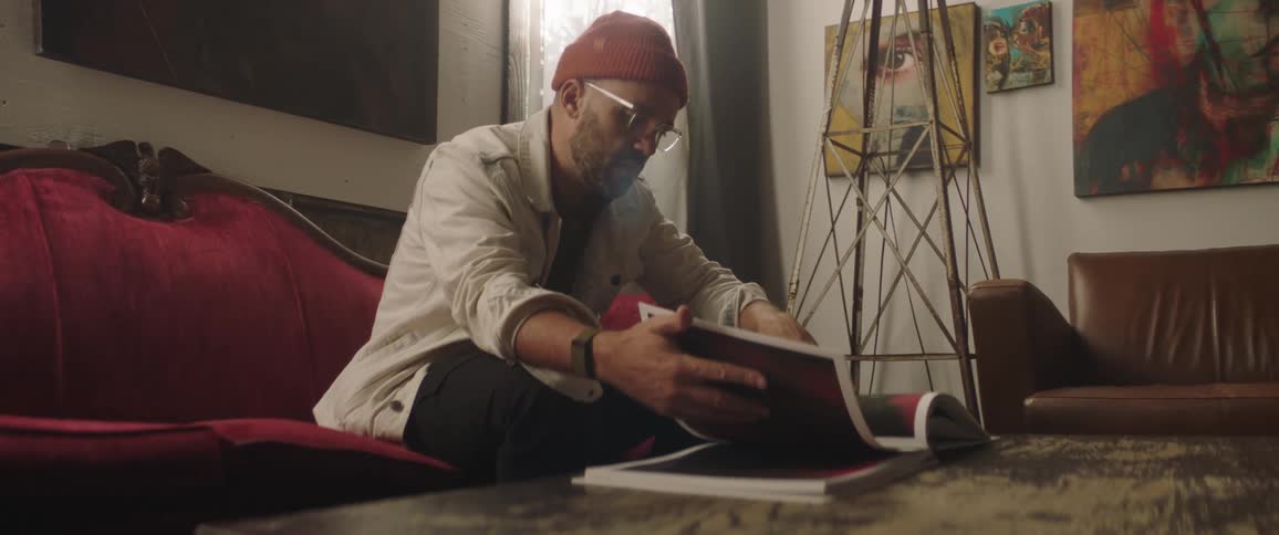 A man with a hat in a room sitting on a couch looking through a book with a wall and a window in the background