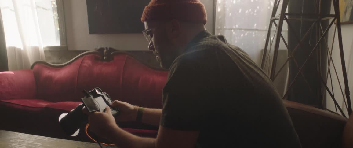 A man operates the buttons on the back of a camera while sitting in the living room of a house during the day