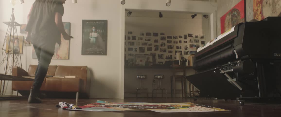 A man with a hat in a room kneeling in front of a canvas with a couch in the background