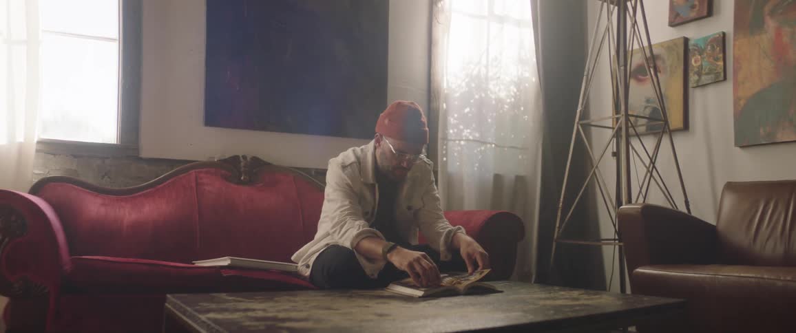 A man sitting on a red couch in a living room flipping through a book on top of a coffee table during the day