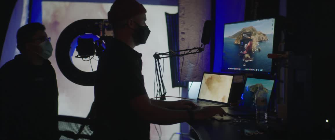 Photographer in studio works with computer showing images as another man watches both wear face masks