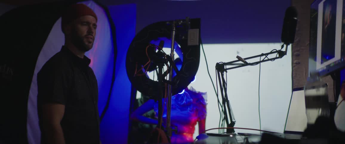 A man in a studio with a camera and a microphone with a woman and a screen in the background
