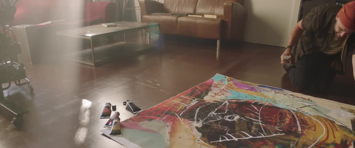 A man with a hat in a room applying paint to a canvas on the floor