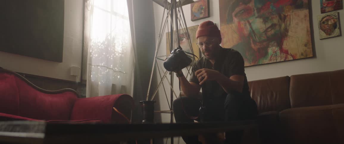 A male photographer sits on a couch and aims his camera at a cup on a coffee table