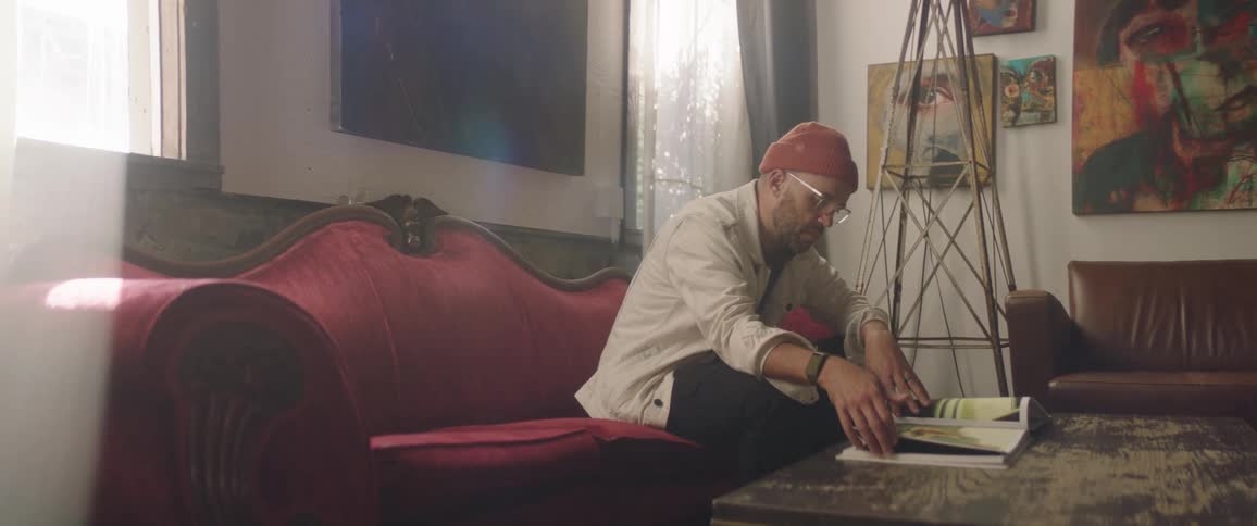 Man wearing a toque and eyeglasses sits on a red sofa and looks through an album of artwork