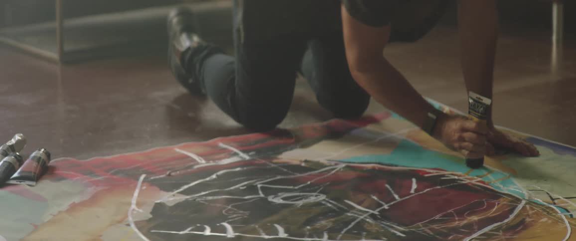 Woman with a red woolly hat a beard and a dark shirt paints a large paper with tubes in the art studio