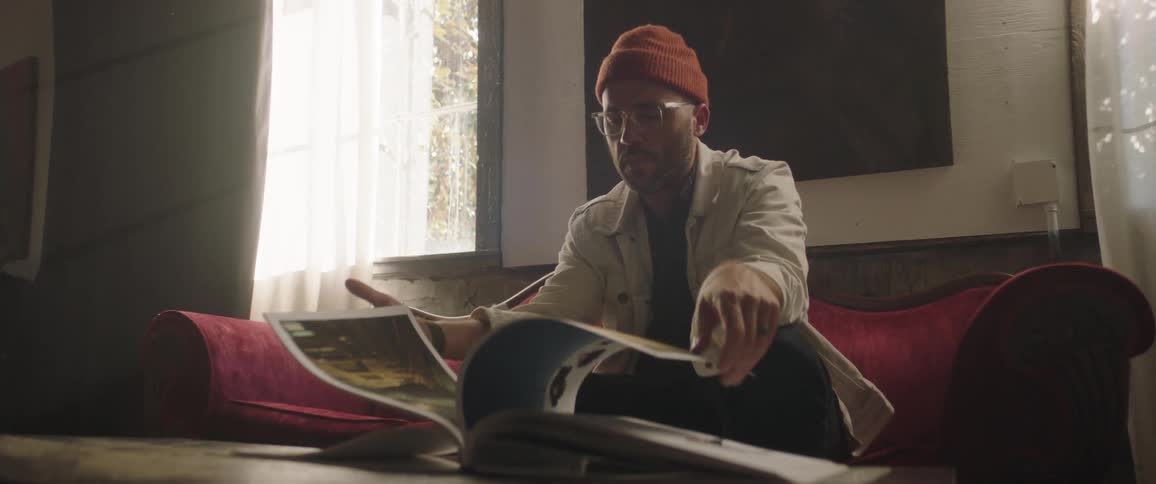 Man with red woolly hat glasses beard and white jacket sits on red couch and turns the pages of magazine