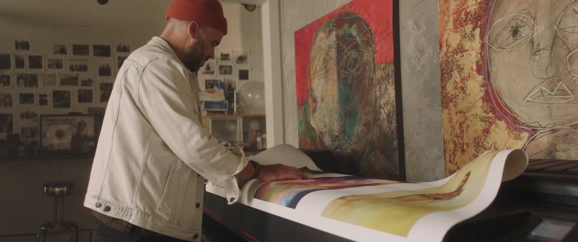Artist stands looking at large printout of art near large scale printer near wall decorated with paintings