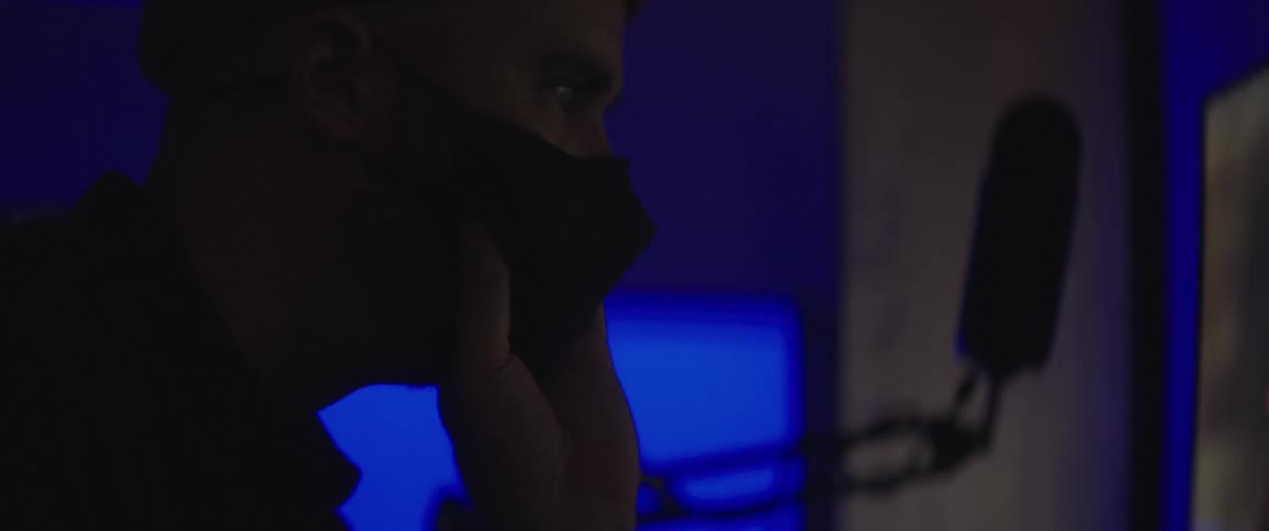 A masked man uses a computer at a desk in a dark room