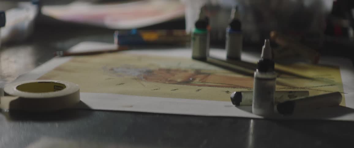Paint bottles sit on sheet of art also covered by spool of tape and pencil