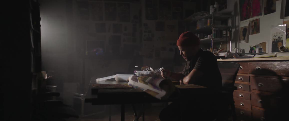 Artist sitting on a desk painting with oils over wood inside a dimly lit studio