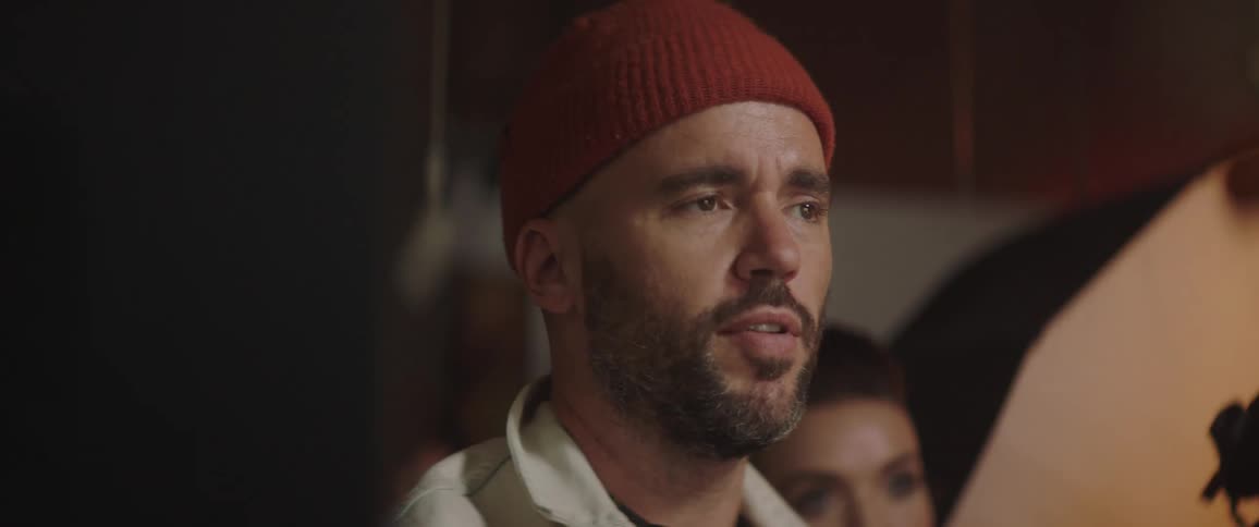 Man wearing a toque raises his arm and talks while a woman stands behind him