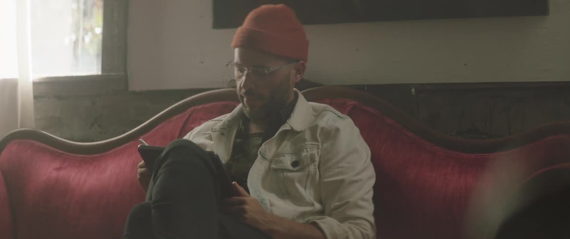 Man wearing glasses and a toque sits on a red sofa and uses a stylus on a tablet