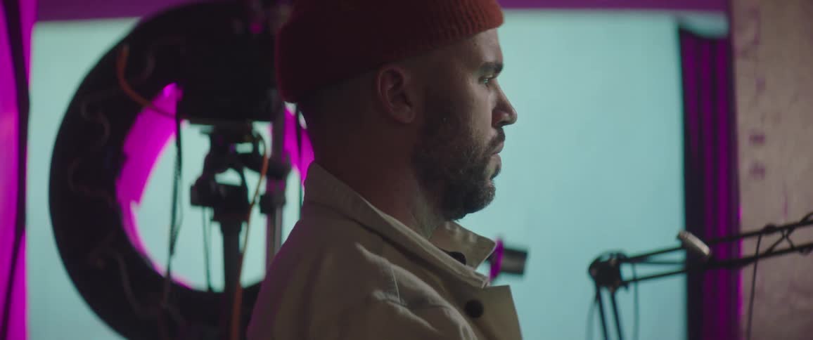 A man with a hat in a studio with a microphone and a screen in the background