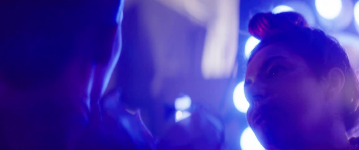 A makeup artist applies makeup to a performer with a brush while bright lights shine behind her