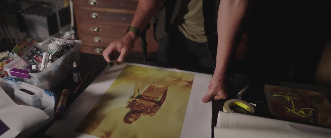 Man wearing a toque stands at a table and works on a piece of art