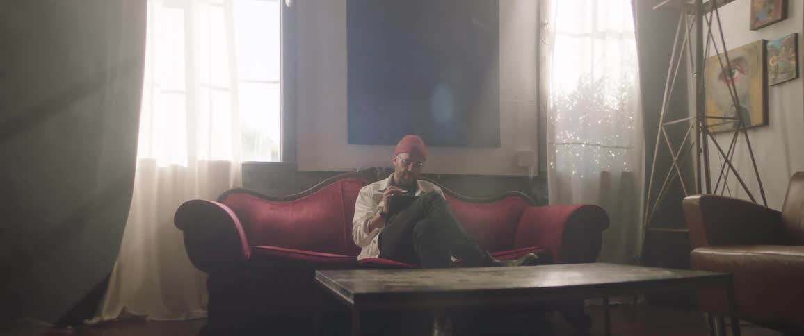A man sits on a red couch in a room writing in a journal during the day