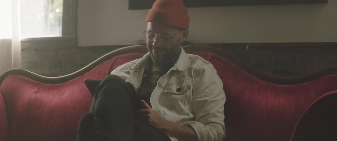 A man with a hat in a room sitting on a couch with a wall and a window in the background