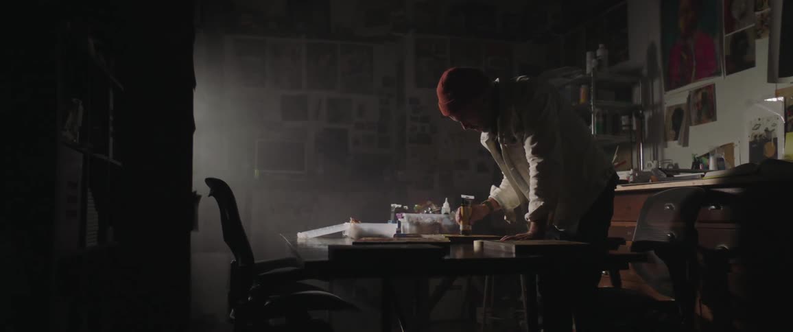 A man with a hat standing by a table and applying paint on a canvas